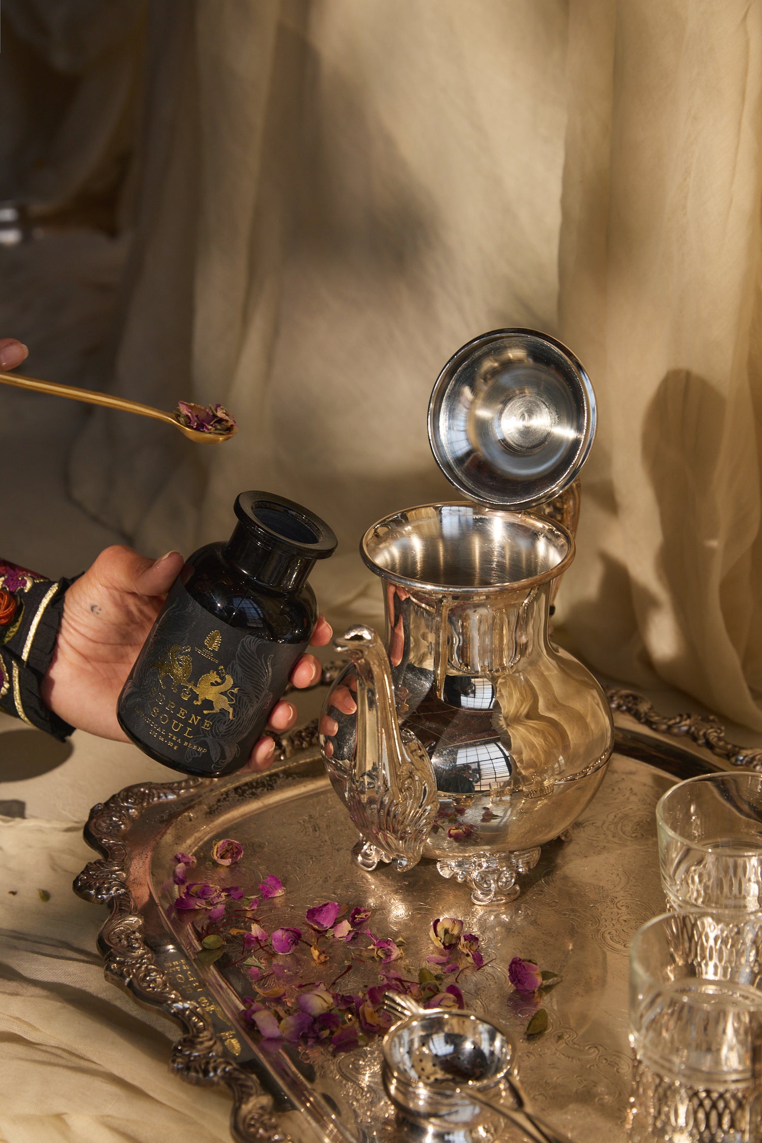 Silver teapot and black container on a tray with a person's hand holding a spoon.